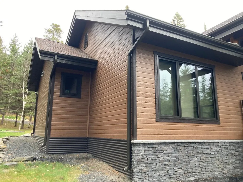 Two-storey home in brown wood-look siding with black seamless eavestrough, black fascia and a black downspout over a stone veneer base